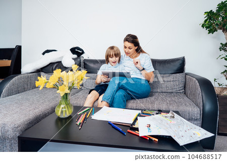 Mother and son on the sofa using digital tablet. Happy mom and little boy using touch screen tablet for watching videos, reading books, playing video game, using online app, studying together at home Mother and son on the sofa using digital tablet. Happy mom and little boy using touch screen tablet for watching videos, reading books, playing video game, using online app, studying together at home 104688517