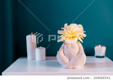 Close up white table with ceramic female body-shaped vase with rose flower, burning candles on turquoise blue wall background. Zen, cozy, meditation female bedroom decor. Self recovery place at home. Close up white table with ceramic female body-shaped vase with rose flower, burning candles on turquoise blue wall background. Zen, cozy, meditation female bedroom decor. Self recovery place at home. 104688541