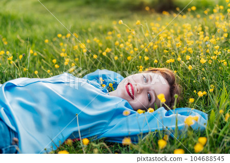 Portrait of smiling happy young woman in blue blouse laying on green grass lawn with yellow flowers. Female enjoy the moment, relaxing outdoors at sunset. Springtime, mental health, no allergy concept Portrait of smiling happy young woman in blue blouse laying on green grass lawn with yellow flowers. Female enjoy the moment, relaxing outdoors at sunset. Springtime, mental health, no allergy concept 104688545