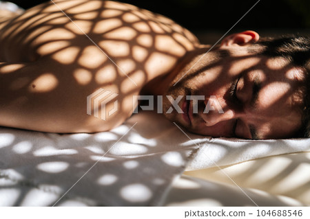 Close up face of sleeping handsome man relaxing lying down with closed eyes on massage table in sunlight and beautiful shadows in spa salon. Concept of body care and rehabilitation of spiritual peace Close up face of sleeping handsome man relaxing lying down with closed eyes on massage table in sunlight and beautiful shadows in spa salon. Concept of body care and rehabilitation of spiritual peace 104688546