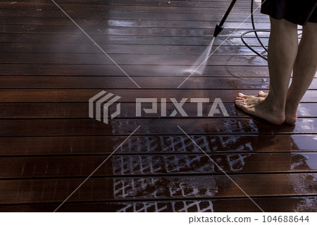 Power surface washing. Man cleaning terrace with a power washer. High water pressure cleaner on wooden terrace surface. 104688644