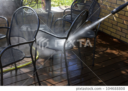 Power surface washing. Man cleaning terrace with a power washer. High water pressure cleaner on wooden terrace surface and outdoor furniture. Power surface washing. Man cleaning terrace with a power washer. High water pressure cleaner on wooden terrace surface and outdoor furniture. 104688808