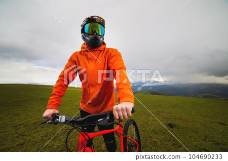 Portrait of a male downhill cyclist in a full face helmet and goggles with an orange windbreaker jacket with a raincoat Portrait of a male downhill cyclist in a full face helmet and goggles with an orange windbreaker jacket with a raincoat 104690233