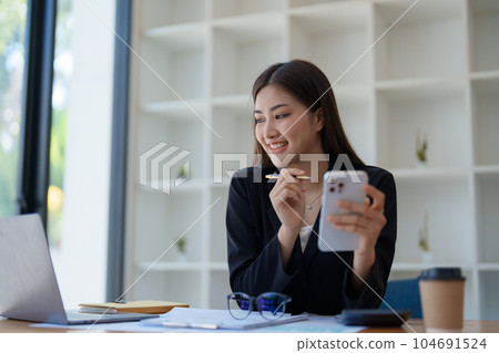 Portrait of a young Asian woman showing a smiling face as she uses her phone, computer and financial documents on her desk in the early morning hours. 104691524