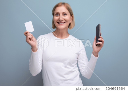 blond middle aged woman in white sweater holding money card mockup and smartphone 104691664