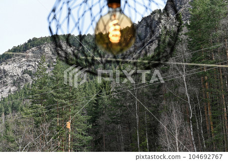 Ceiling chandelier with a mesh frame hangs against the backdrop of high mountains with trees in the background. nice view from the window Ceiling chandelier with a mesh frame hangs against the backdrop of high mountains with trees in the background. nice view from the window 104692767