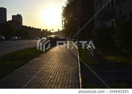 Summer city street in the setting sun. People walking away into the distance. City summer road and sidewalk. 104692855