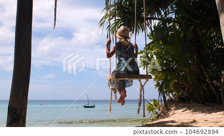 Little girl in straw hat and blue sundress enjoying ocean landscape while swinging on tropical beach Little girl in straw hat and blue sundress enjoying ocean landscape while swinging on tropical beach 104692894