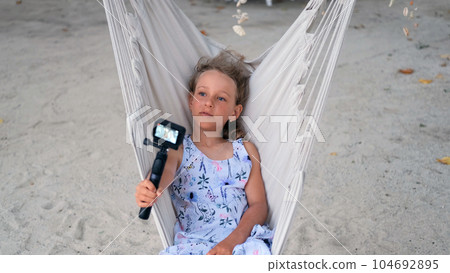 Little girl blogger video streaming from beach while swinging on hammock. Child recording travel vlog Little girl blogger video streaming from beach while swinging on hammock. Child recording travel vlog 104692895