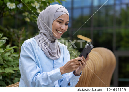 Young beautiful muslim woman in hijab uses phone outside office building, smiling woman reads online news, browses internet pages and dials message call. Young beautiful muslim woman in hijab uses phone outside office building, smiling woman reads online news, browses internet pages and dials message call. 104693129