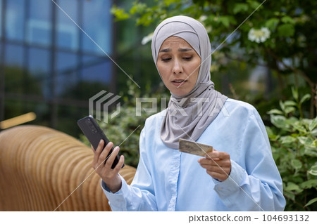 Upset cheated woman with phone trying to make online purchase and money transfer, muslim woman in hijab disappointed got rejection, holding phone and bank credit card, dissatisfied buyer. 104693132