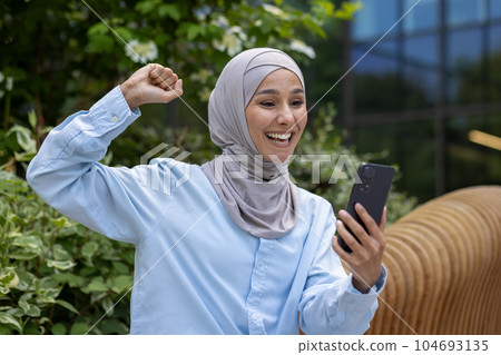 Successful joyful muslim woman in hijab received online win notification, Dinka reading news and holding hand up triumph gesture celebrating sitting on bench outside office building in park. 104693135