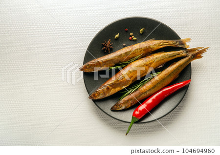 Smoked capelin on a dark plate with rosemary and pepper is served for lunch. Light space for advertising 104694960