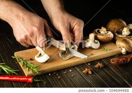 Slicing mushrooms with a knife by the hands of a cook on a cutting board. Cooking mushrooms on the kitchen table with spices and rosemary 104694988