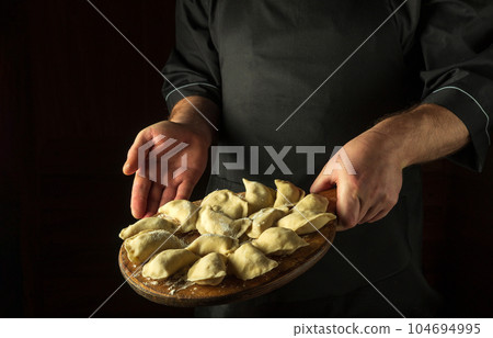 The chef prepares dumplings in the kitchen. Cutting board with raw dumplings in the cook hand. Copy space The chef prepares dumplings in the kitchen. Cutting board with raw dumplings in the cook hand. Copy space 104694995