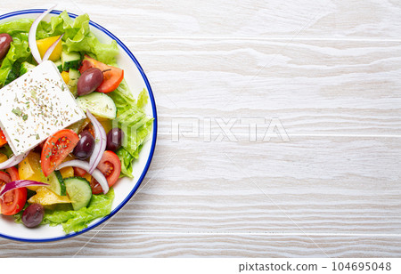 Traditional Greek Salad with Feta Cheese, Tomatoes, Bell Pepper, Cucumbers, Olives, Herbs in white ceramic bowl on White rustic wooden table background from above, Cuisine of Greece. Space for text Traditional Greek Salad with Feta Cheese, Tomatoes, Bell Pepper, Cucumbers, Olives, Herbs in white ceramic bowl on White rustic wooden table background from above, Cuisine of Greece. Space for text 104695048