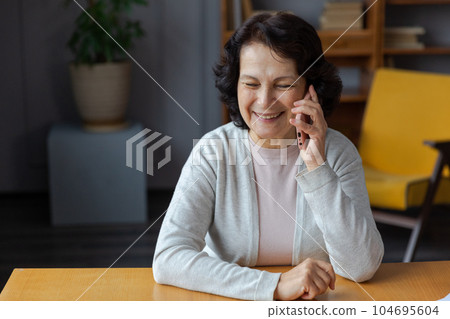 Happy middle aged senior woman talking on smartphone with family friends. Older mature lady with cell phone chatting with grown up children, resting in at home. Older generation modern tech usage 104695604