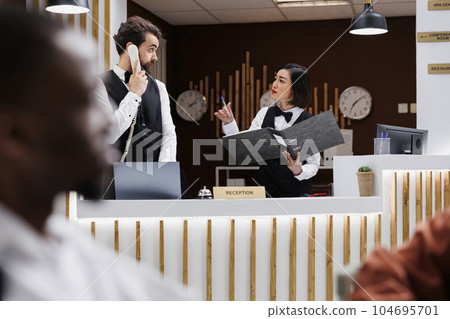 Diverse reception staff multitasking at front desk, working on online room bookings with landline phone and welcoming guests at arrival. Team of receptionists checking hotel records. 104695701