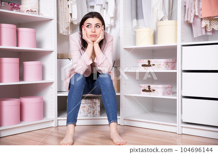 A young beautiful brunette woman in jeans and a pink jacket sits in a dressing room with her face in her hands meditating A young beautiful brunette woman in jeans and a pink jacket sits in a dressing room with her face in her hands meditating 104696144