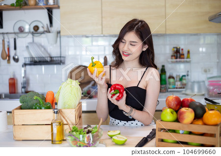 beautiful asian woman smiling preparing, holding a sweet pepper and cut green apple in white kitchen with plenty of fresh vegetables and fruits on the table. At Home 104699660