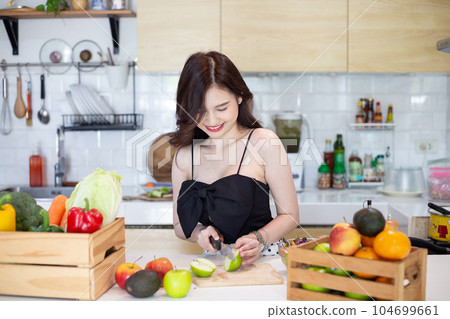 beautiful asian woman smiling preparing, cut green apple in white kitchen with plenty of fresh vegetables and fruits on the table. 104699661