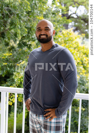 Portrait of happy biracial man in pyjamas standing on sunny balcony at home in front of treetops 104700144