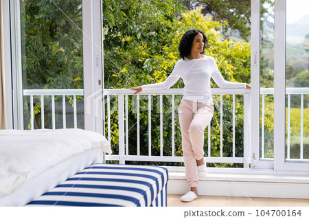 Happy biracial woman in slippers relaxing at balcony in bedroom with treetops in background 104700164