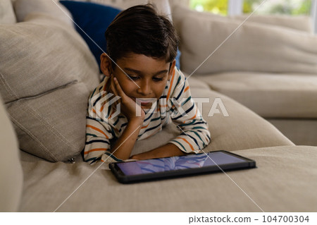 Happy biracial boy lying on couch using tablet with copy space in living room 104700304