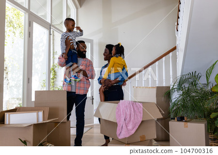 African american parents carrying children while standing by cardboard boxes in new home 104700316