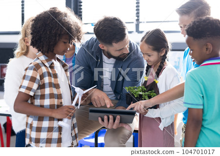 Diverse elementary schoolchildren and male teacher using wind turbine and tablet in classroom 104701017