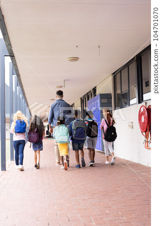 Vertical rear view of diverse male teacher and children in elementary school corridor, copy space Vertical rear view of diverse male teacher and children in elementary school corridor, copy space 104701070