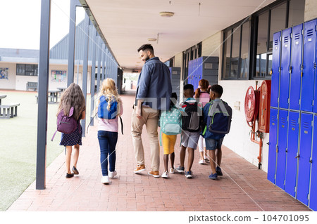 Rear view of diverse male teacher and children walking in elementary school corridor, copy space Rear view of diverse male teacher and children walking in elementary school corridor, copy space 104701095