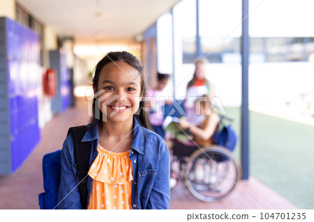 Portrait of smiling biracial schoolgirl standing with bag in school corridor 104701235