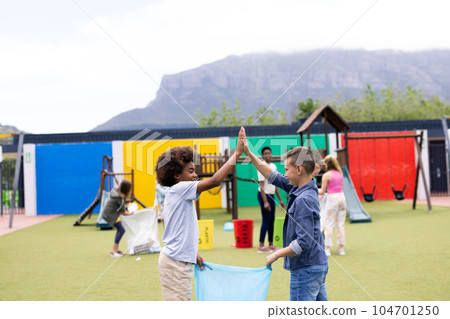 Two diverse schoolboys collecting rubbish for recycling high fiving in schoolyard, with copy space 104701250