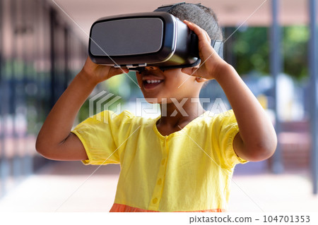 Happy biracial schoolgirl using vr headset in sunny school corridor Happy biracial schoolgirl using vr headset in sunny school corridor 104701353