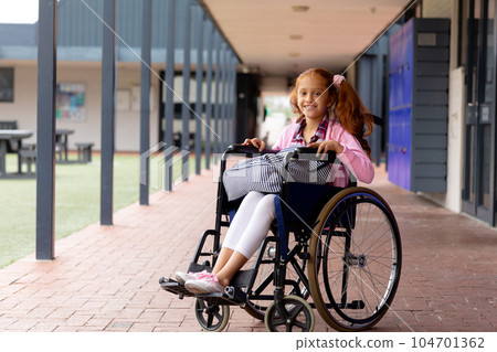 Portrait of happy biracial schoolgirl in wheelchair, smiling in school corridor 104701362