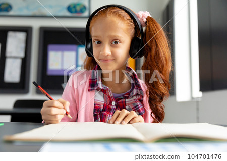 Portrait of happy biracial schoolgirl sitting at desk wearing headphones in class with copy space 104701476