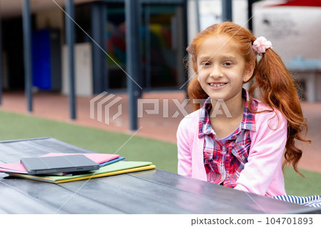 Portrait of happy biracial schoolgirl at table with books and tablet in schoolyard 104701893