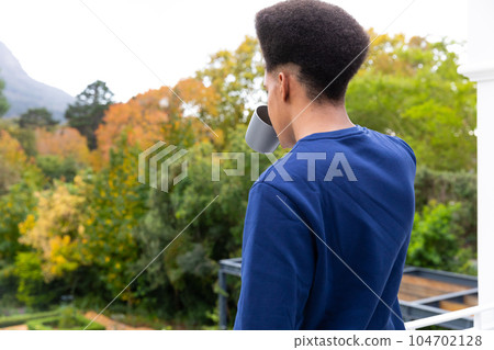 Rear view of thoughtful biracial man standing on balcony drinking coffee and enjoying view of garden 104702128