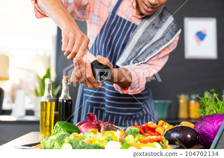 Midsection of biracial man wearing apron cooking dinner, seasoning vegetables in kitchen Midsection of biracial man wearing apron cooking dinner, seasoning vegetables in kitchen 104702134