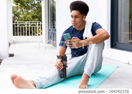 Biracial man using smartphone and earphones drinking water sitting on sunny balcony after yoga 104702178