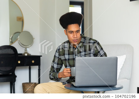 Biracial man sitting at home having cup of coffee and using laptop 104702196