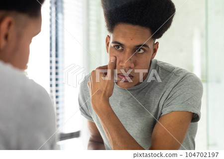 Biracial man looking in mirror inspecting face and eyes in sunny bathroom Biracial man looking in mirror inspecting face and eyes in sunny bathroom 104702197