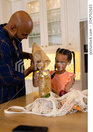 Happy biracial father and daughter pouring pasta from paper bag into storage jar in kitchen Happy biracial father and daughter pouring pasta from paper bag into storage jar in kitchen 104702683