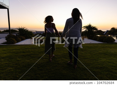 Biracial lesbian couple holding hands and walking in garden at sunset Biracial lesbian couple holding hands and walking in garden at sunset 104702684
