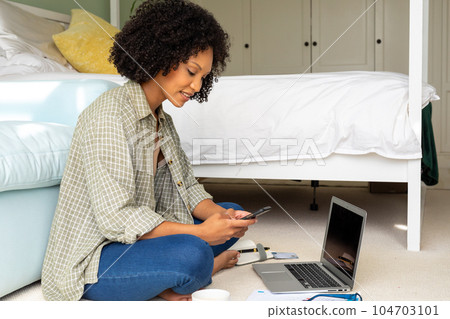 Happy biracial woman using laptop and smartphone sitting on floor in bedroom 104703101