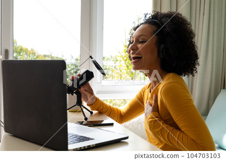 Happy biracial woman using headphones, laptop and microphone podcasting from home 104703103