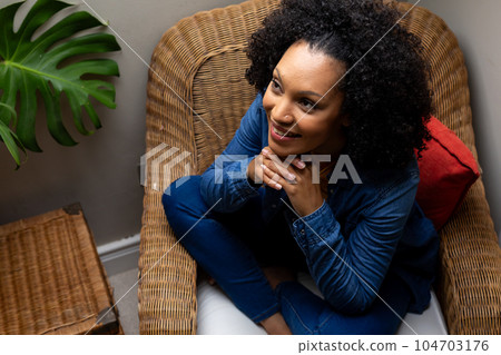 Happy biracial woman sitting on woven armchair at home Happy biracial woman sitting on woven armchair at home 104703176