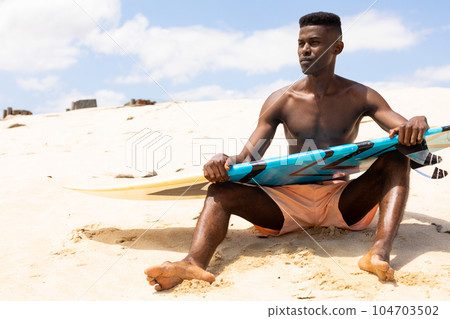 Focused biracial man holding surfboard sitting on sunny beach looking away Focused biracial man holding surfboard sitting on sunny beach looking away 104703502
