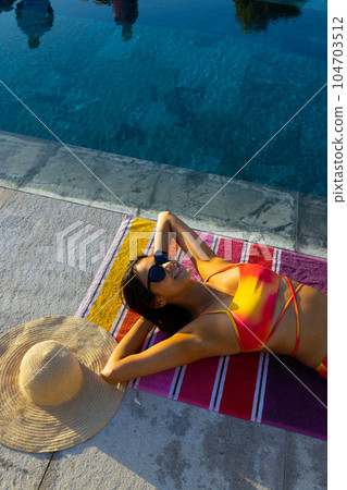 Happy biracial woman on towel sunbathing next to swimming pool in garden, copy space 104703512